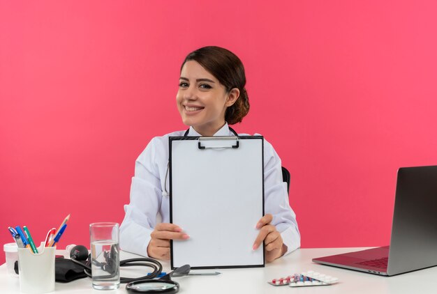 Happy young female doctor wearing medical robe and stethoscope sitting at desk with medical tools and laptop showing clipboard isolated