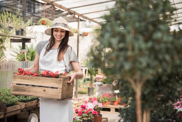 温室に立つベゴニアの花の木箱を持つ幸せな若い女性の庭師