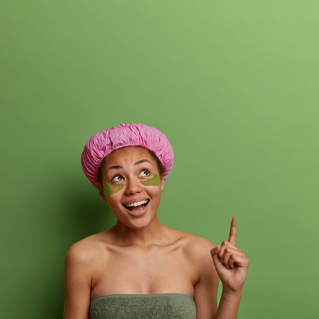 Happy young female model points above with fore finger, recommends cosmetology product for healthy skin, wears bath hat and green patches under eyes, feels positive, poses indoor, empty space up