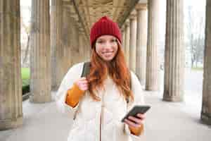 Free photo happy young female model stands on street and holds mobile phone uses smartphone app follows the rou