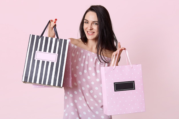 Free photo happy young female shopaholic with pleased facial expression, dressed in summer polka dot dress, holds shopping bags, rejoices buying new clothes, poses on rosy. woman with packages