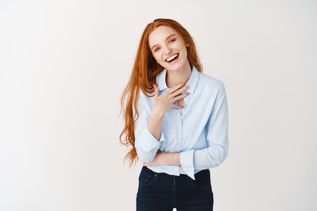 Happy young female with long red hair pointing at herself, laughing and looking confident, standing over white wall