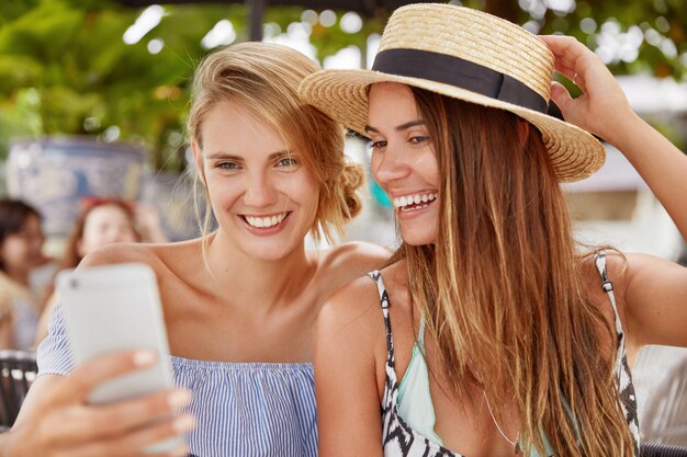幸せな若い女性は、スマートフォンで面白いビデオを見たり、自分撮りをしたり、楽しそうな顔をしたり、リゾート都市の屋外カフェテリアで一緒に休んだりしています。人、関係、夏の残りのコンセプト