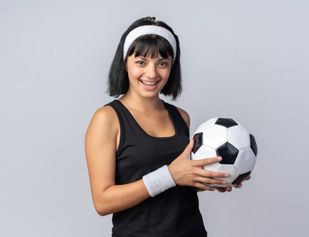 Happy young fitness girl wearing headband holding soccer ball looking at camera smiling cheerfully