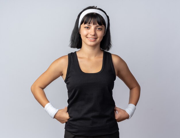 Happy young fitness girl wearing headband looking at camera smiling confident standing over white