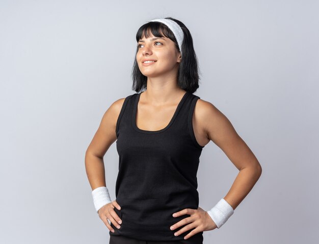 Happy young fitness girl wearing headband looking up smiling cheerfully standing over white background