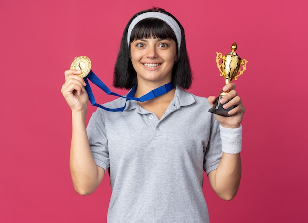 Happy young fitness girl wearing headband with gold medal around neck holding trophy