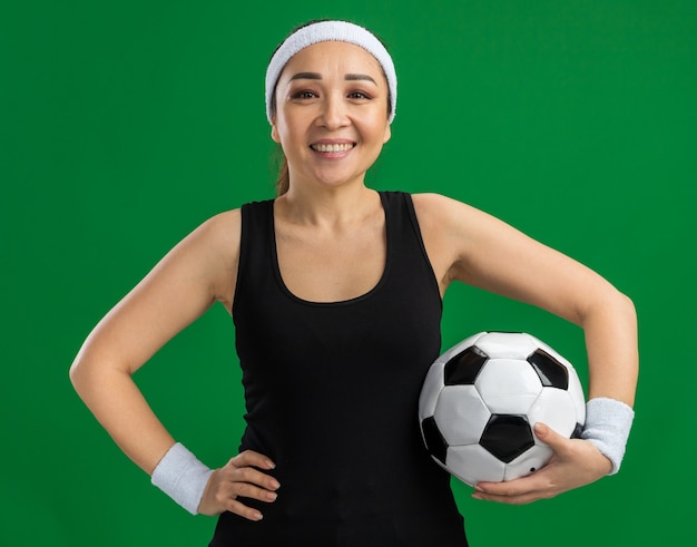 Foto gratuita felice giovane donna fitness con fascia e bracciali che tengono un pallone da calcio con un sorriso sul viso in piedi sul muro verde