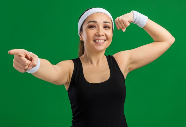 Счастливая молодая фитнес-женщина с повязкой на голову и повязками, улыбаясь, уверенно поднимая кулак, показывая бицепсы
