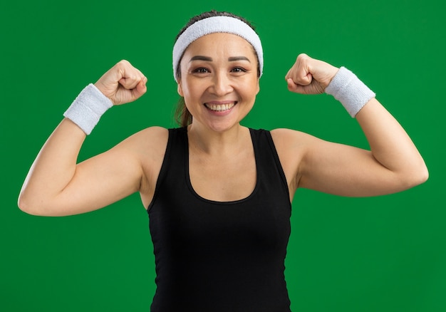 Happy young fitness woman with headband and armbands  smiling confident raising fists standing over green wall