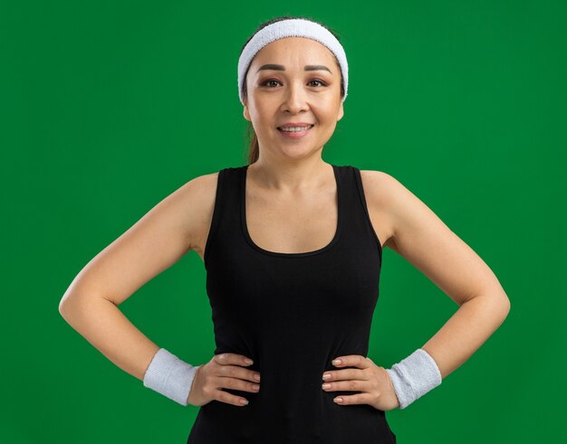 Happy young fitness woman with headband and armbands  smiling confident standing over green wall