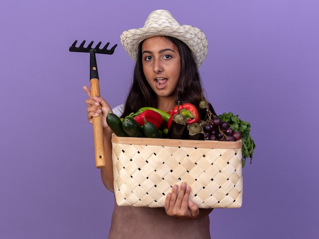 Happy young gardener girl in apron and summer hat holding crate full of vegetables and mini rake  with smile on face standing over purple wall