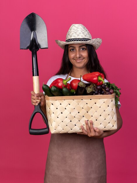 Foto gratuita felice giovane giardiniere ragazza in grembiule e cappello estivo tenendo la cassa piena di verdure e pala con il sorriso sul viso in piedi sopra il muro rosa