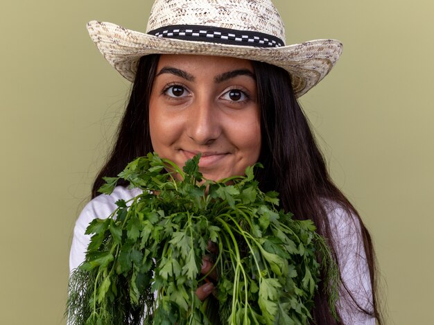 Happy young gardener girl in apron and summer hat holding fresh herbs  with smile on face standing over green wall