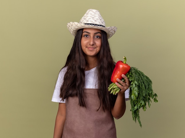 Счастливая молодая девушка-садовник в фартуке и летней шляпе держит свежий красный болгарский перец и свежие травы, улыбаясь, стоя над зеленой стеной