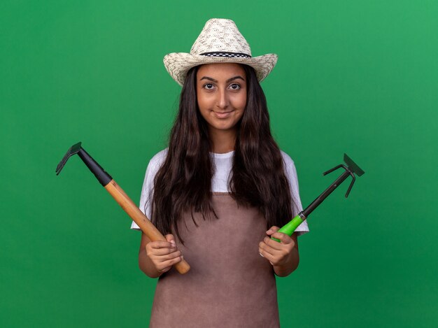 Happy young gardener girl in apron and summer hat holding mini rake and mattock  with smile on face standing over green wall