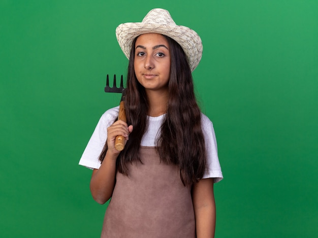 Felice giovane ragazza giardiniere in grembiule e cappello estivo che tiene mini rastrello con il sorriso sul viso in piedi sopra la parete verde