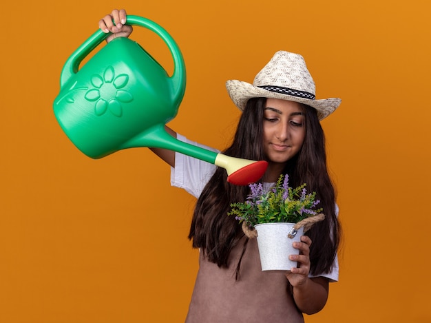 オレンジ色の壁の上に立っている顔に笑顔でじょうろと鉢植えの植物の散水植物を保持しているエプロンと夏の帽子の幸せな若い庭師の女の子