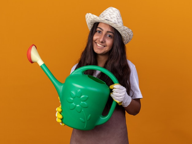 Felice giovane ragazza giardiniere in grembiule e cappello estivo che tiene annaffiatoio con il sorriso sul viso in piedi sopra la parete arancione