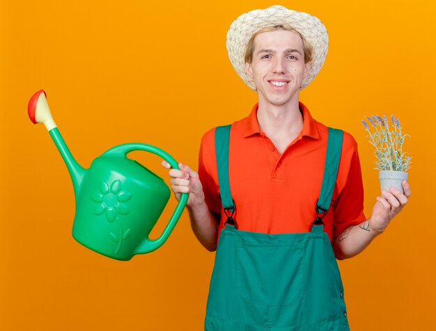 Happy young gardener man wearing jumpsuit and hat holding watering can