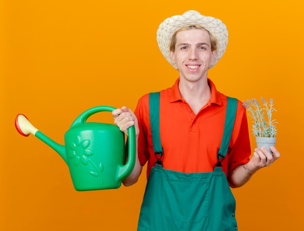 Happy young gardener man wearing jumpsuit and hat holding watering can