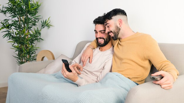 Happy young gay couple sitting together on sofa looking at mobile phone