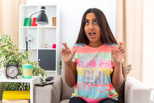 Happy young girl in casual clothes making wish crossing fingers with hope expression sitting on the chair in light living room