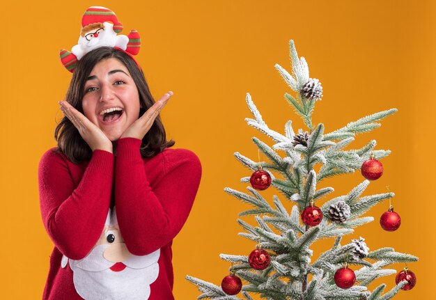 Счастливая молодая девушка в рождественском свитере с забавной повязкой на голову, глядя в камеру, весело улыбаясь, стоя рядом с елкой на оранжевом фоне