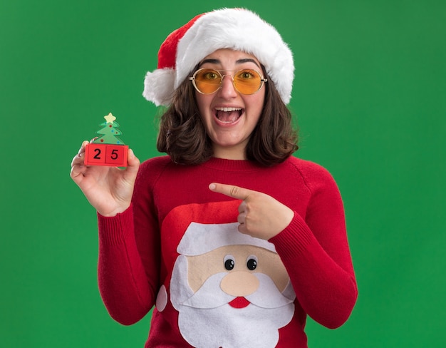 Happy young girl in christmas sweater wearing santa hat and glasses holding toy cubes with number twenty five pointing with index finger at it smiling cheerfully standing over green wall