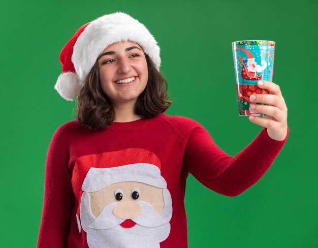 Happy young girl in christmas sweater wearing santa hat holding colorful paper cup looking at it with smile on face standing over green wall