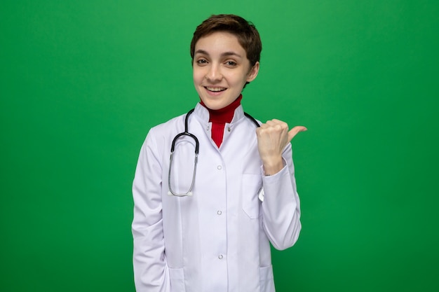 Happy young girl doctor in white coat with stethoscope around neck smiling confident pointing with thumb back standing on green