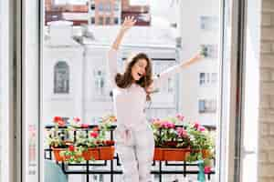 Foto gratuita felice giovane ragazza in pigiama divertendosi sul balcone nella mattina di sole. alza le mani e tiene gli occhi chiusi.