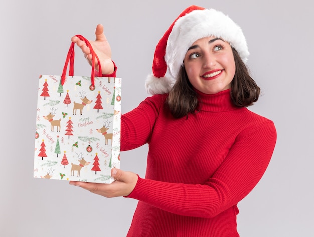 Happy young girl in red sweater and santa hat holding colorful paper bag with christmas gifts looking aside with smile on face standing over white background