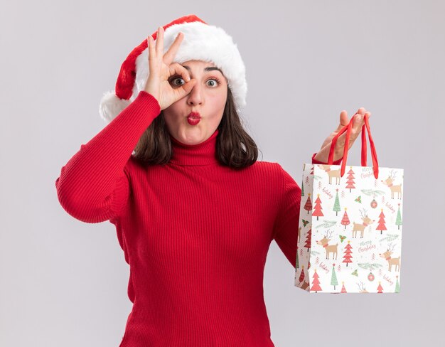 Happy young girl in red sweater and santa hat holding colorful paper bag with christmas gifts making ok sign looking through this sign standing over white background