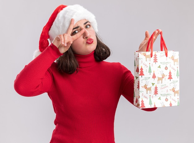 Счастливая молодая девушка в красном свитере и шляпе санта-клауса держит красочный бумажный пакет с рождественскими подарками, показывая знак v рядом с глазом, стоящим над белой стеной
