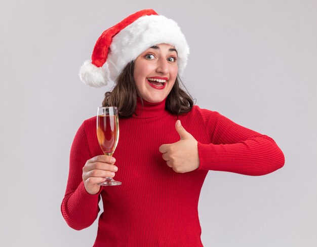 Счастливая молодая девушка в красном свитере и шляпе санта-клауса держит бокал шампанского, глядя в камеру, весело улыбаясь, показывая пальцы вверх, стоя на белом фоне