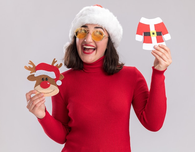 赤いセーターと白い壁の上に立っている顔に笑顔で脇を見てクリスマスのおもちゃを保持している眼鏡をかけているサンタ帽子の幸せな少女