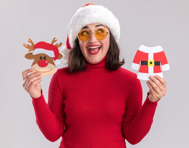 Счастливая молодая девушка в красном свитере и шляпе санта-клауса в очках с рождественскими игрушками, глядя вверх с улыбкой на лице, стоя над белой стеной