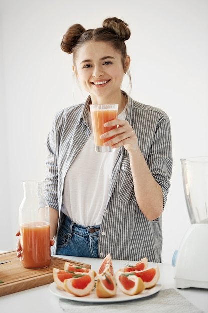 Счастливая маленькая девочка усмехаясь держащ стекло с здоровым smoothie грейпфрута вытрезвителя над белой стеной.