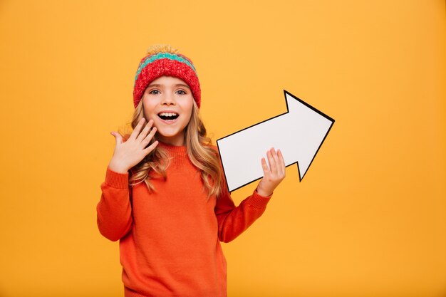 Happy Young girl in sweater and hat pointing with paper arrow away and looking at the camera over orange