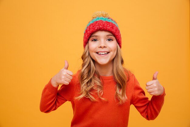 스웨터와 모자 엄지 손가락을 보여주는 오렌지 위로 카메라를 찾고 행복 한 어린 소녀
