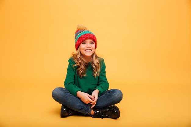 Happy Young girl in sweater and hat sitting on the floor while looking away over orange