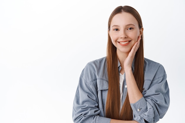 Happy young girl touching her face, smiling pleased, showing smooth gentle facial skin, standig against white wall in cool shirt.