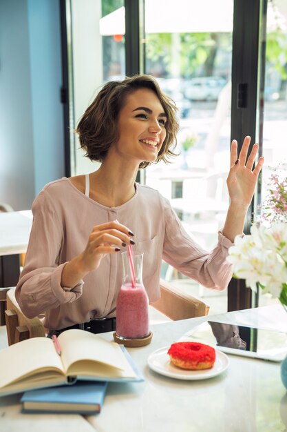 Happy young girl waving hand while drinking smoothie