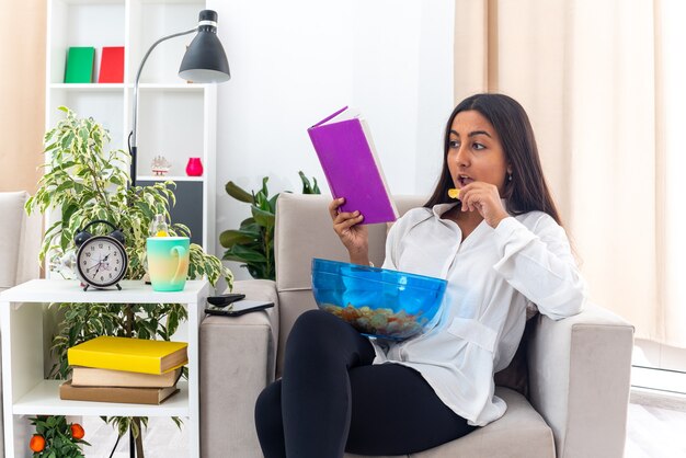 Happy young girl in white shirt and black pants with bowl of chips holding book reading and eating sitting on the chair in light living room