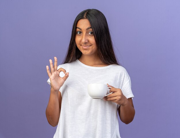 Happy young girl in white t-shirt holding a cup looking at camera smiling showing ok sign standing over blue