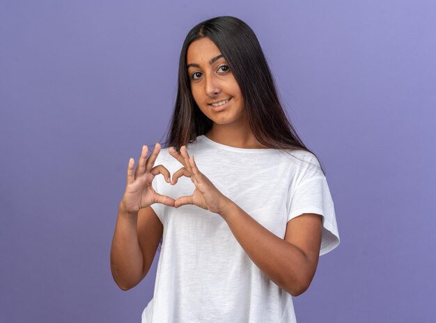 Happy young girl in white t-shirt looking at camera smiling making heart gesture with fingers standing over blue background