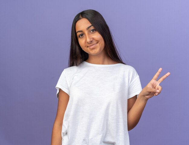 Happy young girl in white t-shirt looking at camera with smile on face showing v-sign standing over blue background