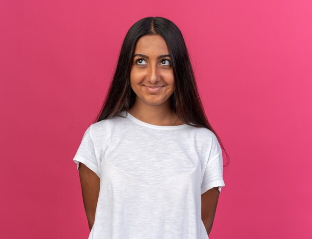 Happy young girl in white t-shirt looking up with shy smile on face standing over pink background