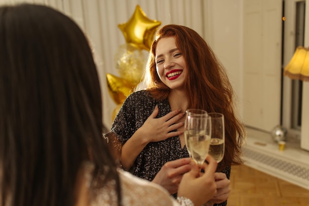 Happy young girls in dresses raising glasses toasting redhead friend's birthday in room Concept of holiday emotions